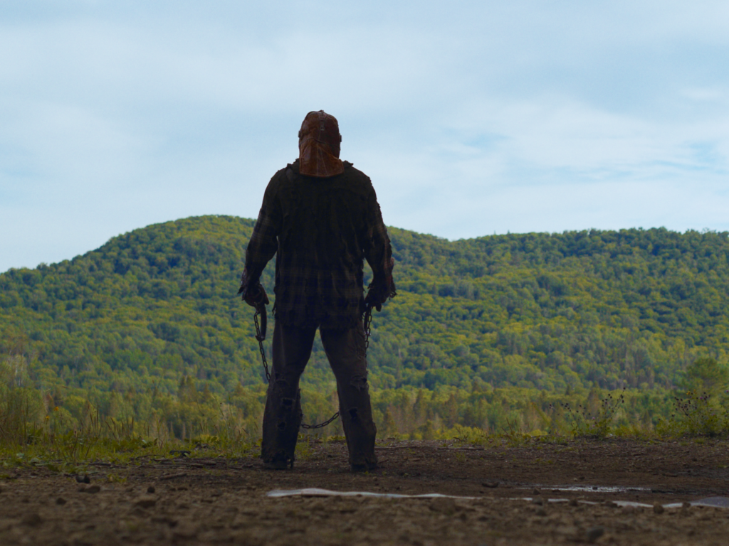 Still from In A Violent Nature showing a large, hooded man holding chains stands on a mountainside, gazing out at a neighbouring mountain. He does not look friendly.
