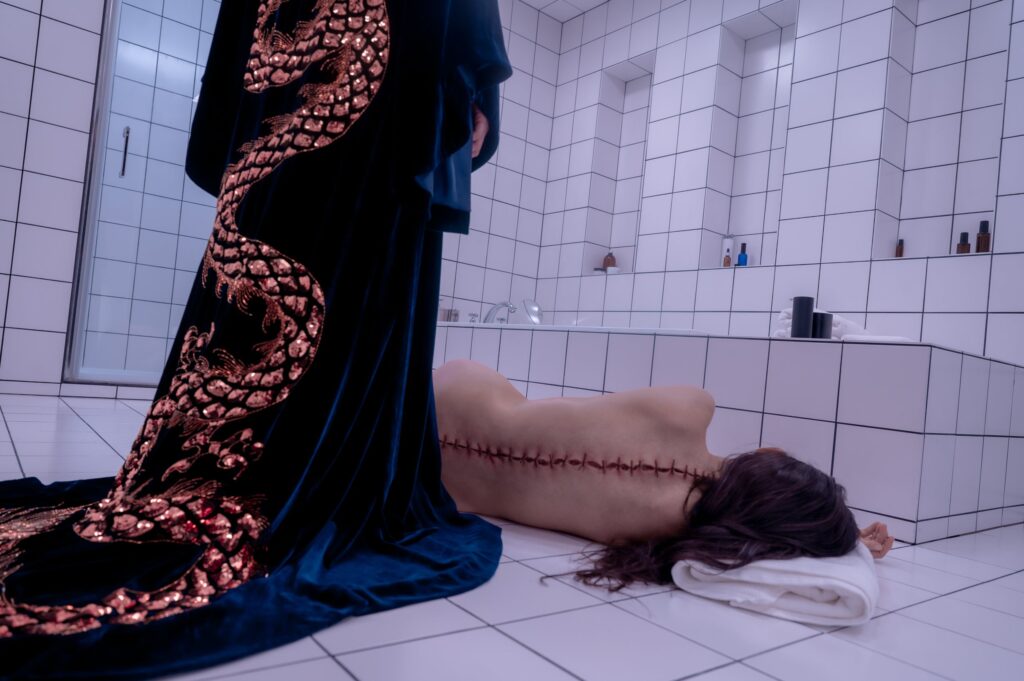 A graphic image of a naked woman laying on a starkly tiled white bathroom, the split skin of her spine roughly stitched together. A figure in a rich velvet robe embroidered with a winding dragon stands over her.