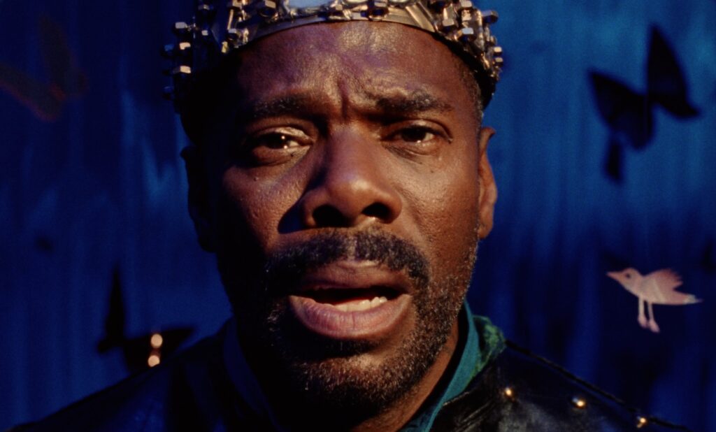 A closeup of a man (Colman Domingo) set against a blue backdrop. He wears a makeshift golden crown and stares ahead, mouth slightly agape.